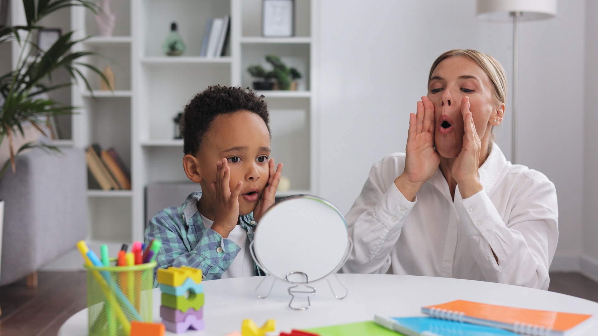 Cute little multiracial boy with speech problems practicing with occupational therapist, practicing and looking at mirror in office. The concept of childhood autism.